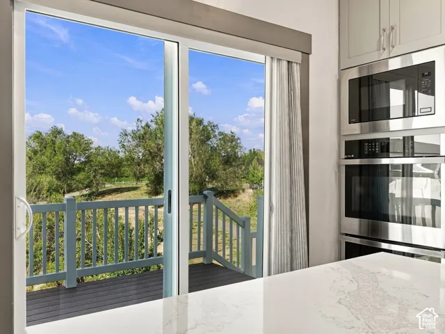 Sliding glass door opening to an elevated deck with wood-finish decking and railing, accompanied by built-in stainless steel double ovens and microwave unit, and light-toned stone-surfaced countertop