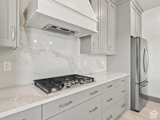 Kitchen featuring gray shaker-style cabinetry, a stone-patterned backsplash, quartz countertops, a stainless steel gas cooktop with a custom range hood, and a stainless steel refrigerator