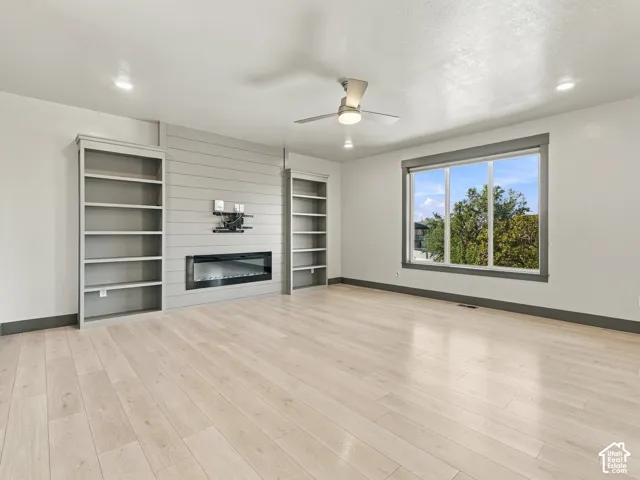 Light-toned wood-finish flooring, linear electric fireplace set in a shiplap feature wall, built-in shelving, large picture window, and ceiling-mounted fan