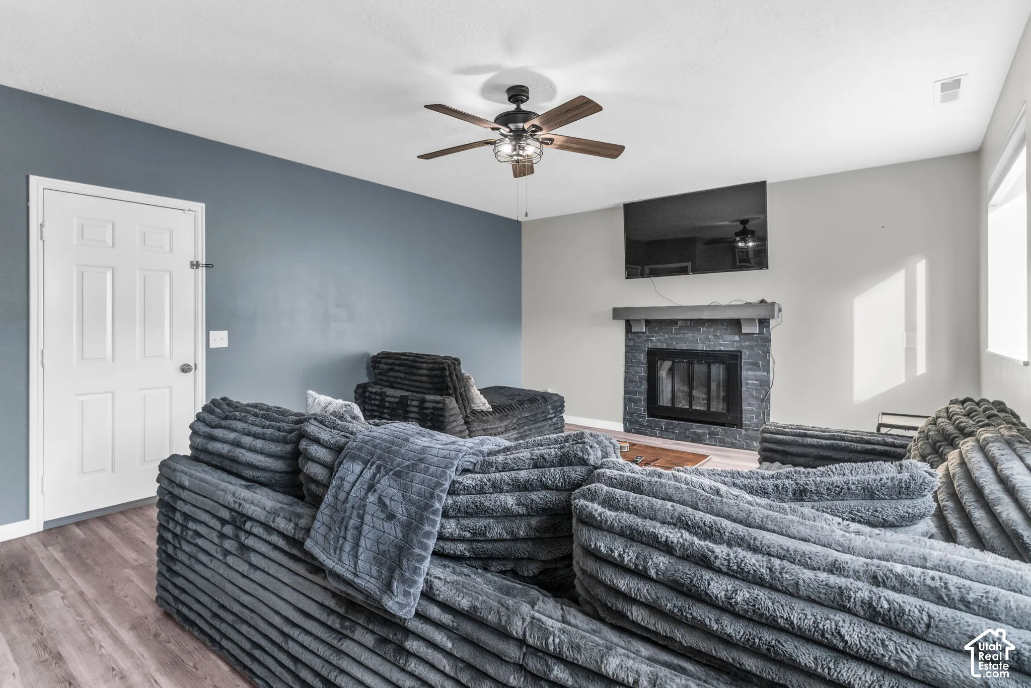 Living area featuring wood-finish flooring, a stacked-stone fireplace with a wooden mantel, an accent wall, and a ceiling fan with integrated lighting
