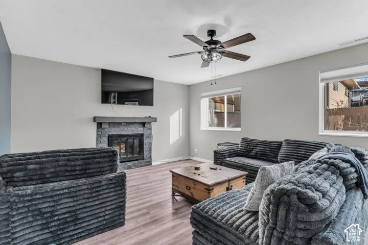 Stone-faced fireplace with a mantel, wood-finish flooring, ceiling fan with integrated lighting, and dual windows