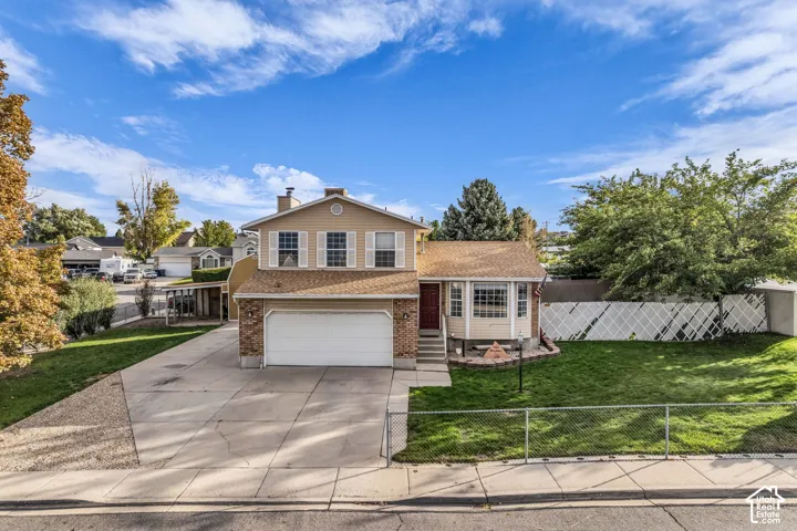 Two-story exterior featuring tan horizontal siding, brick masonry detailing, an attached two-car garage, a front bay window, and a fenced yard with mature landscaping