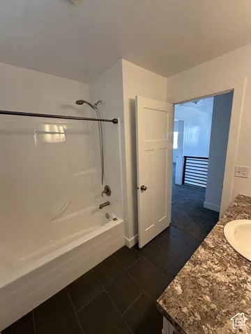 Bathroom featuring a bathtub and shower combination with a wall-mounted handheld sprayer, dark-toned tile flooring, and a stone-patterned vanity countertop