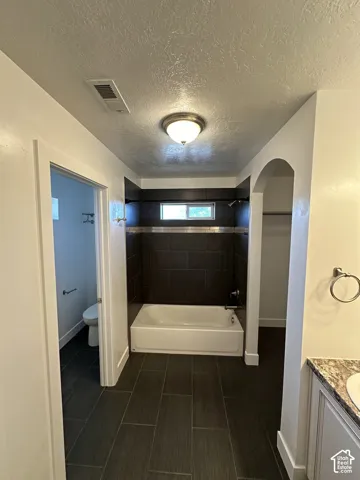 Bathroom featuring large-format dark tile flooring, a soaking tub with a tiled surround, a decorative horizontal mosaic tile accent, and arched architectural detailing