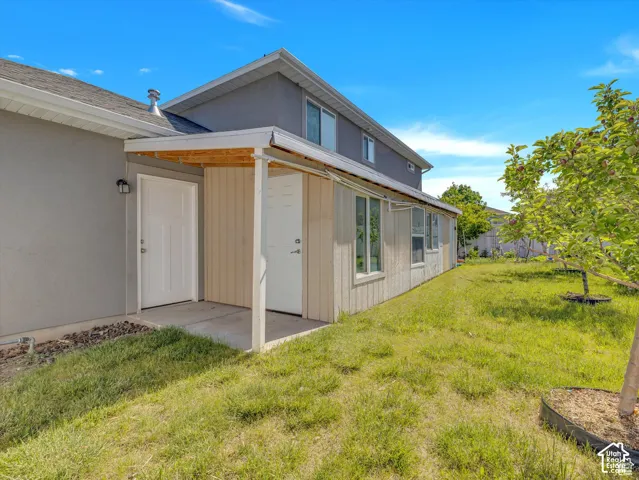 Exterior view featuring a covered patio, neutral-toned stucco and vertical panel siding, and a spacious rear lawn with mature trees