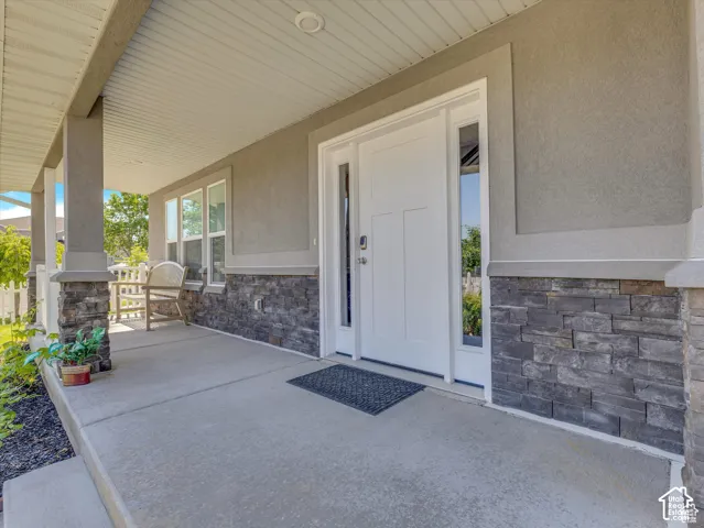 Covered front porch featuring a concrete floor, stone veneer wainscoting, square structural columns with stone bases, and a white front door with dual sidelights