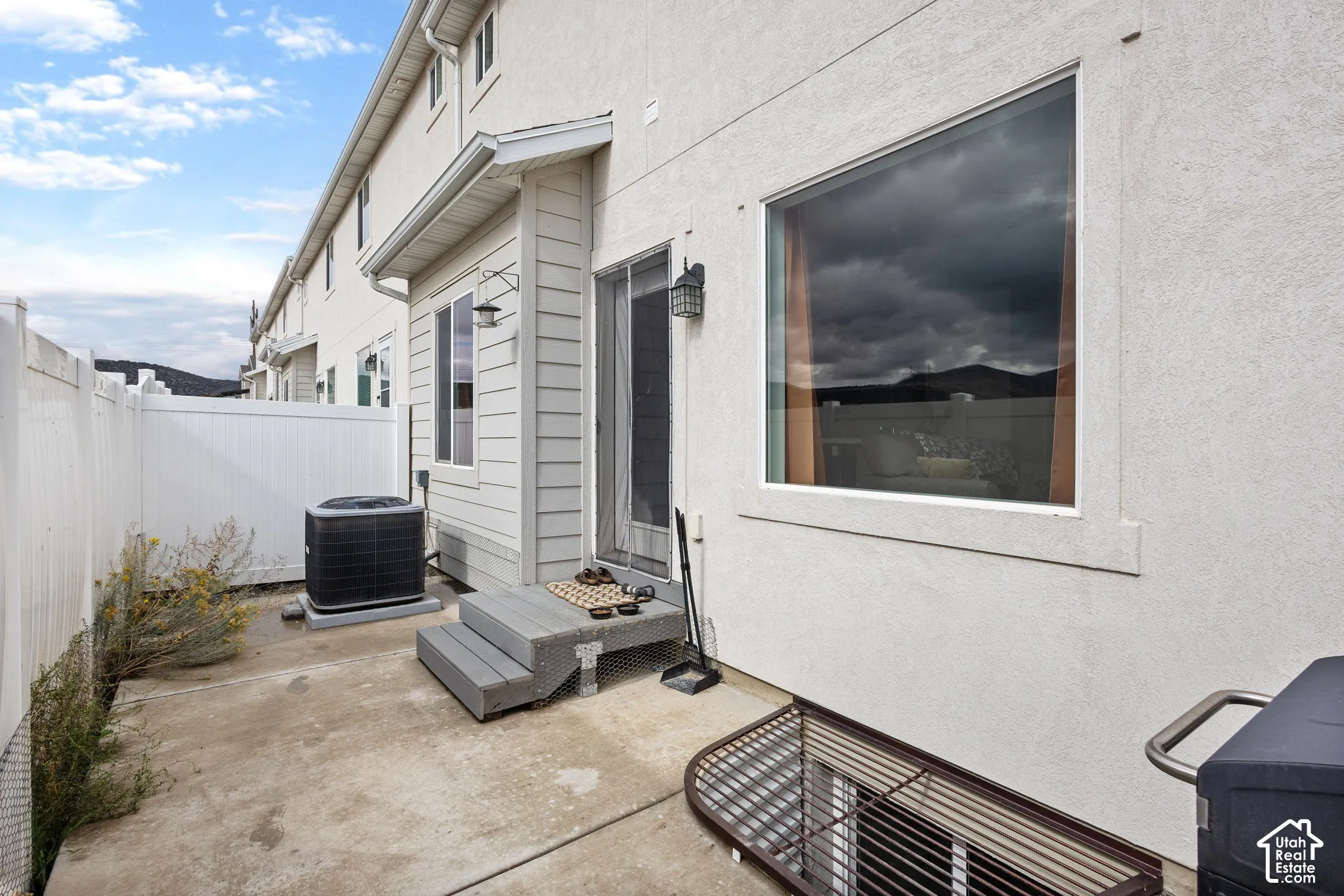 Exterior concrete patio with vinyl privacy fencing, metal window well cover, and wall-mounted outdoor lighting