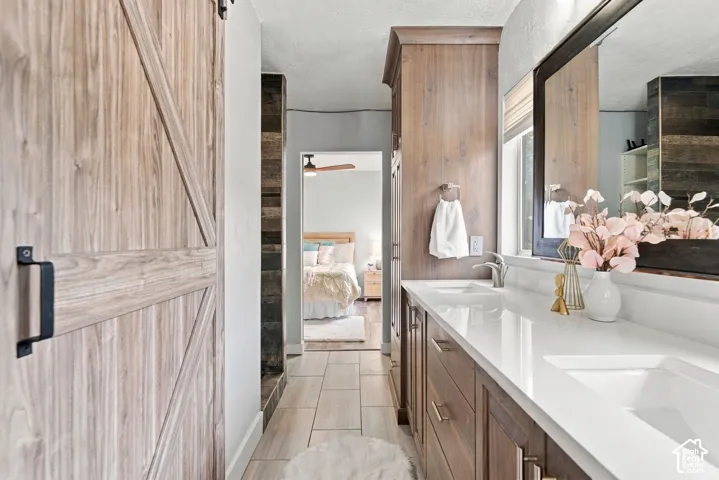 Bathroom featuring a wood-finish barn door, dual-sink vanity with white stone-style countertops, floor-to-ceiling cabinetry, and light-toned tile flooring