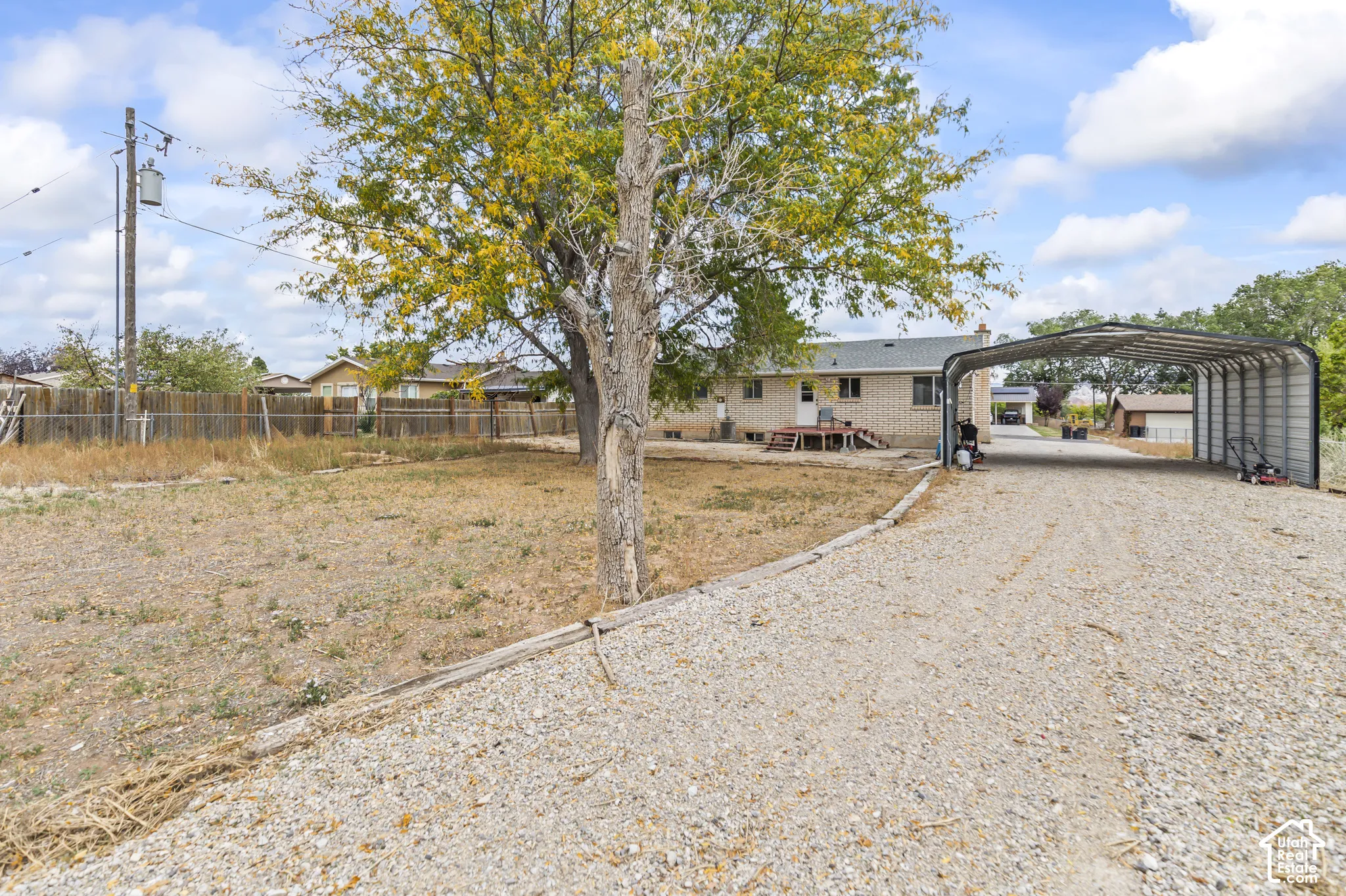 Exterior view featuring a masonry facade, metal-roofed carport, gravel driveway, and wood-slat perimeter fencing