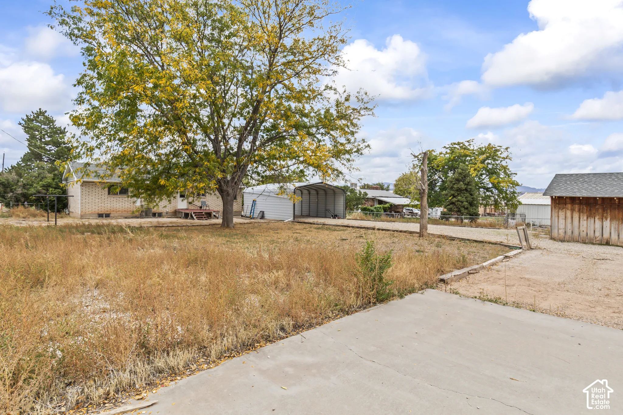 Exterior lot featuring a brick-sided residence, metal carport, wood-sided detached structure, and mature tree