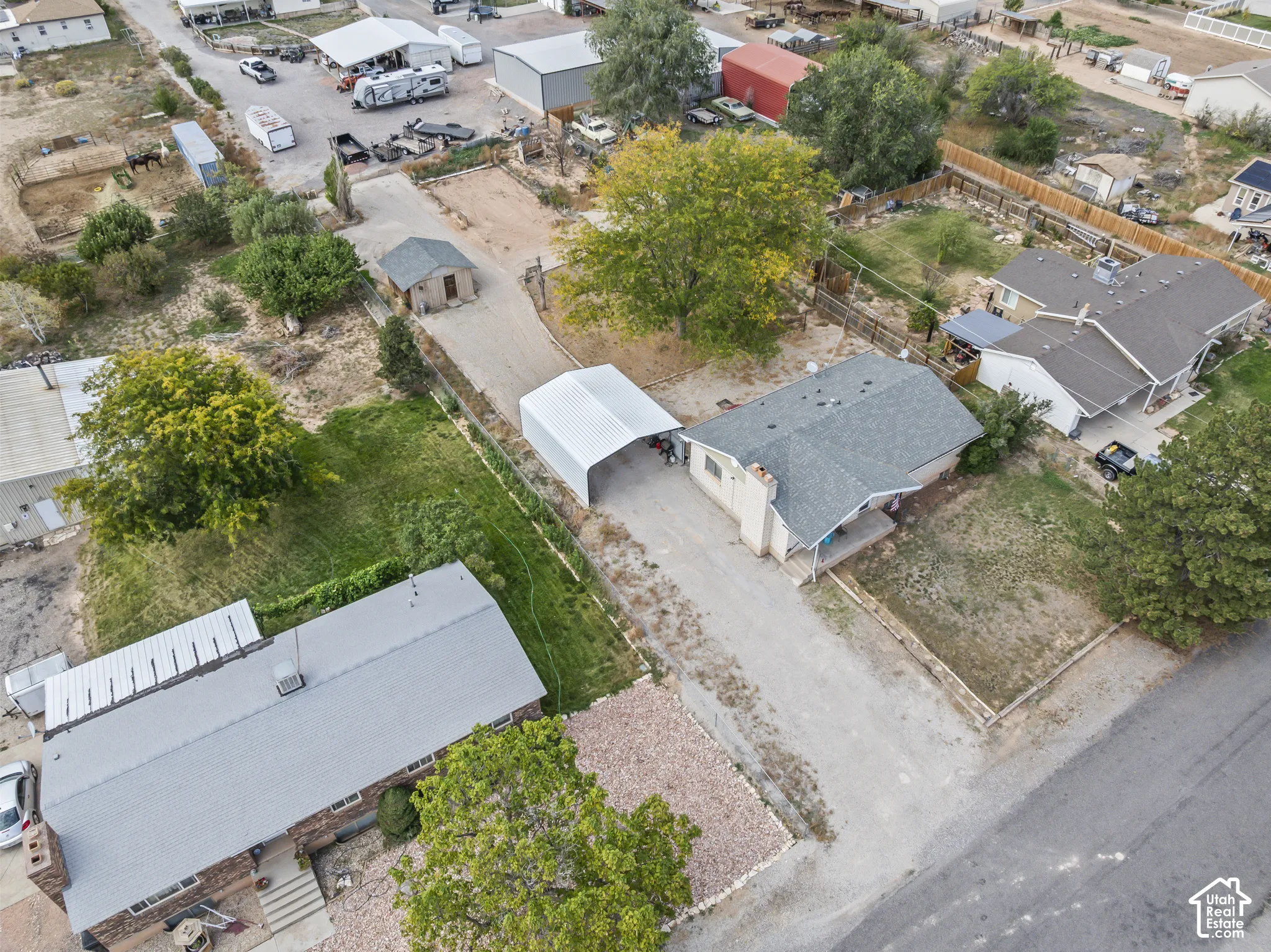 Aerial perspective of a property featuring a single-story residence, a detached metal carport, a separate outbuilding, a gravel driveway, and mature landscaping
