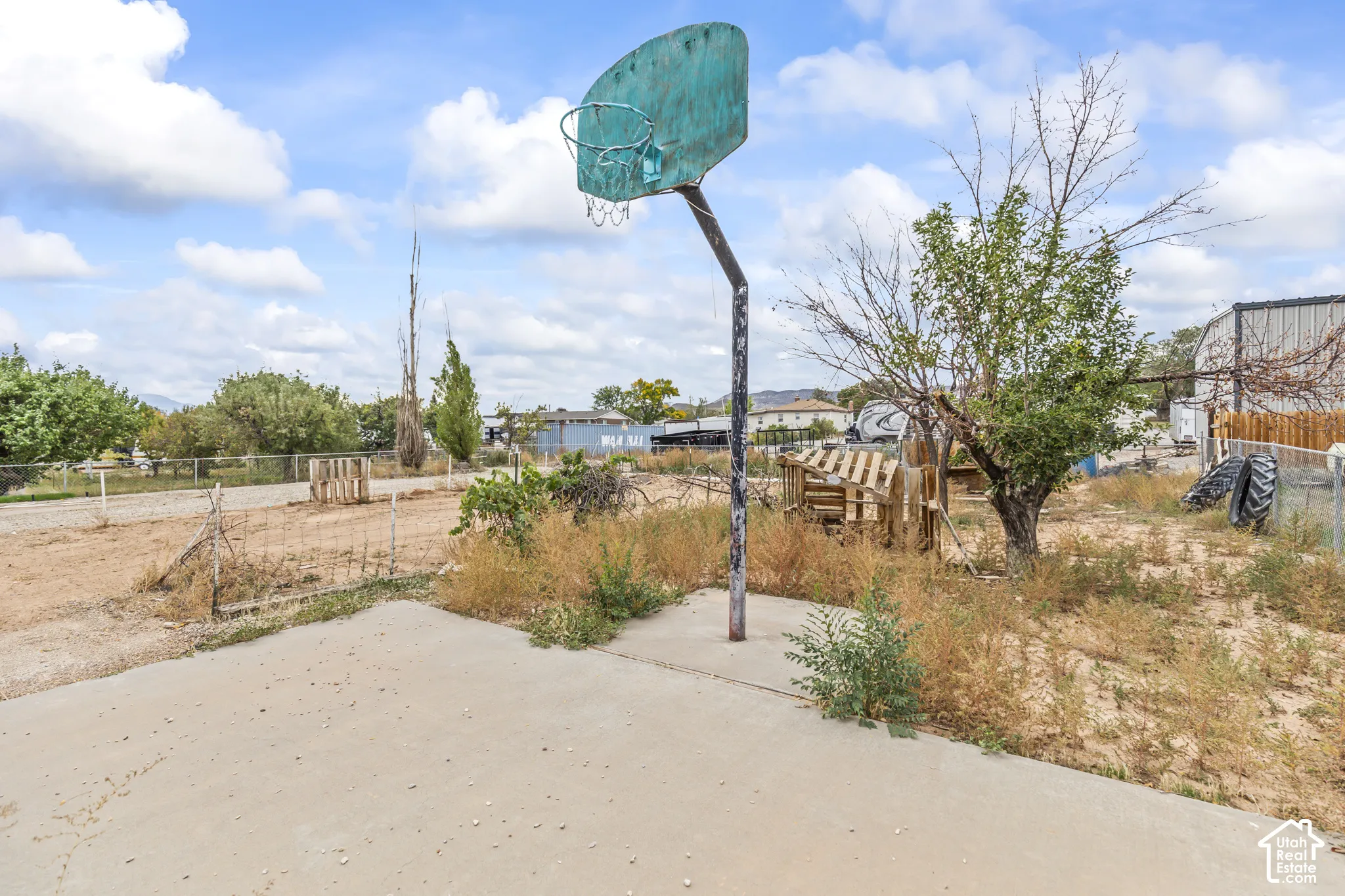 Concrete pad featuring a fixed basketball hoop, chain-link perimeter fencing, and mature trees