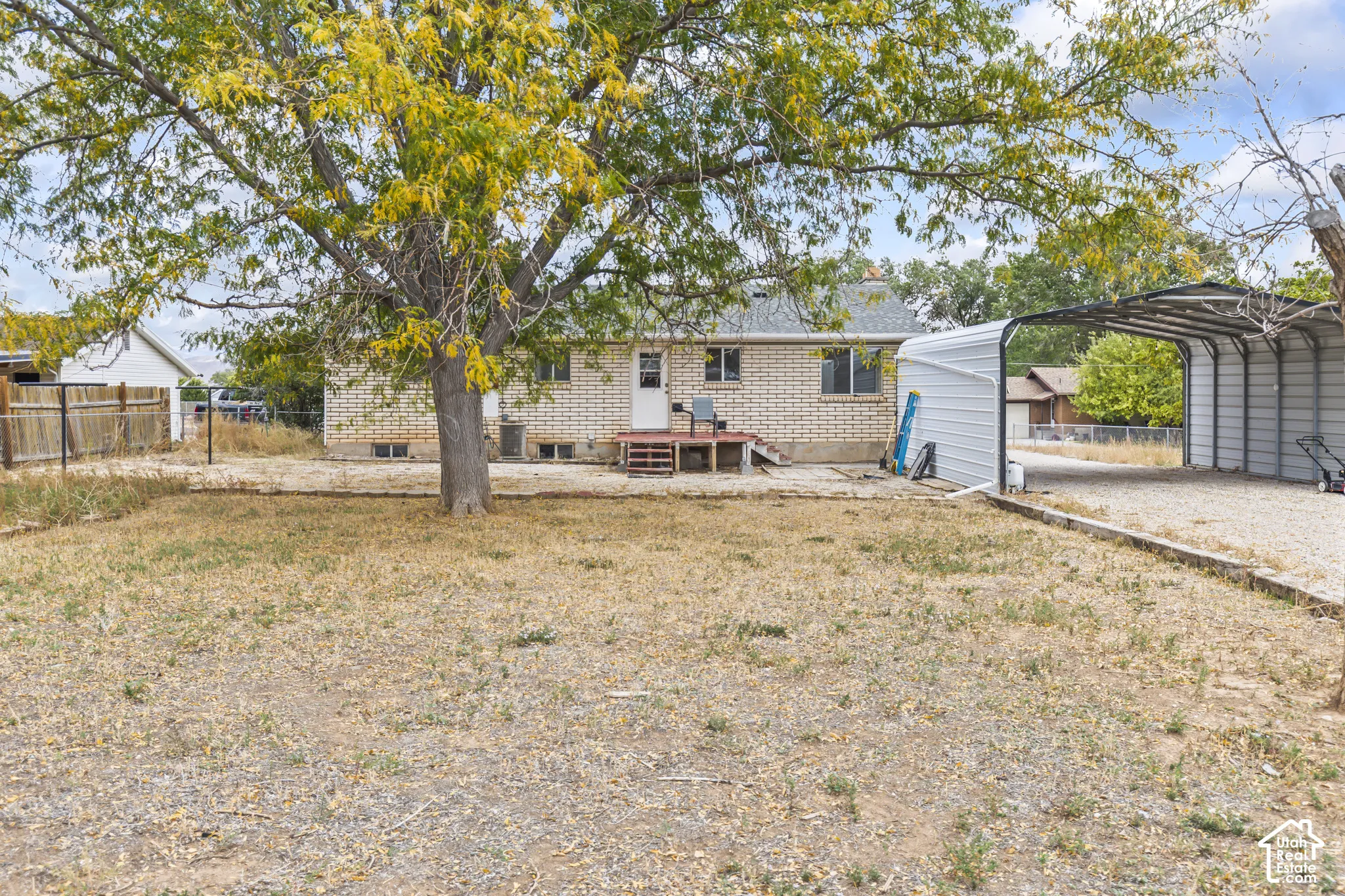 Exterior rear view featuring a brick facade, metal carport, mature tree, and gravel ground cover