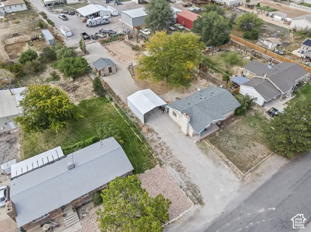Aerial perspective of a property featuring a single-story residence, a detached metal carport, a separate outbuilding, a gravel driveway, and mature landscaping