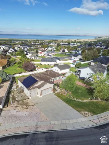 Aerial perspective of a residential property featuring an attached two-car garage, roof-mounted solar panels, concrete driveway, and a fenced yard with mature trees