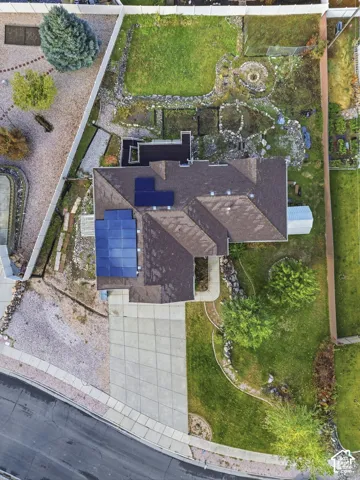 Aerial perspective featuring a roof-mounted solar panel system, concrete driveway, fenced yard, stone-bordered landscaping, and mature trees