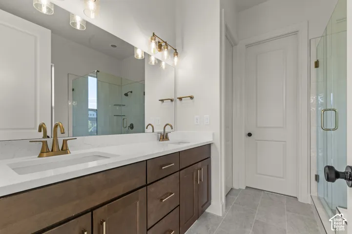 Dual-sink vanity with a quartz countertop, wood-finish cabinetry, brass-toned fixtures, a full-width mirror, and a glass-enclosed shower with tile surround