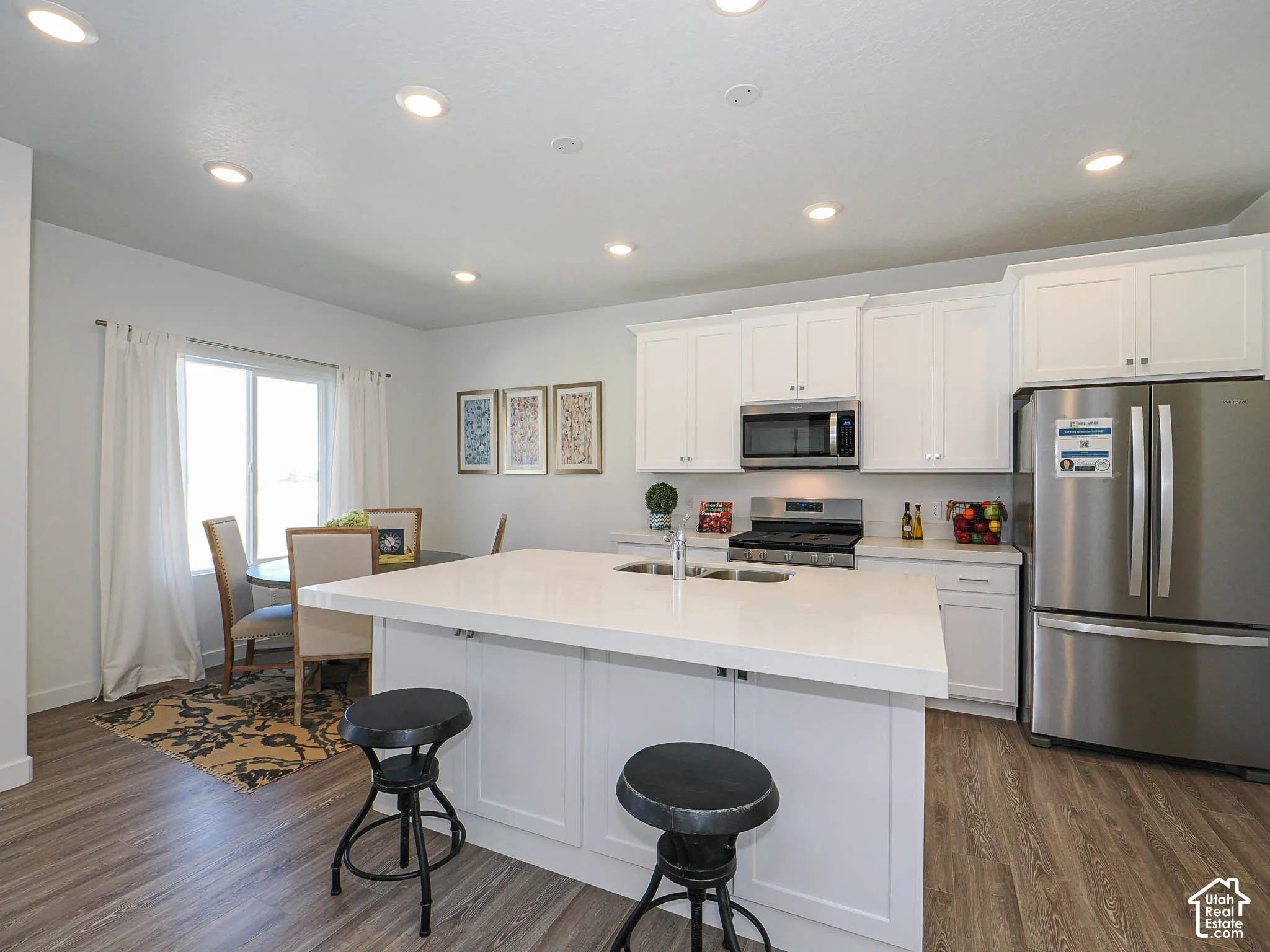 Kitchen featuring white shaker-style cabinetry, quartz countertops, a central island with an integrated sink, stainless steel appliances, and wood-finish flooring
