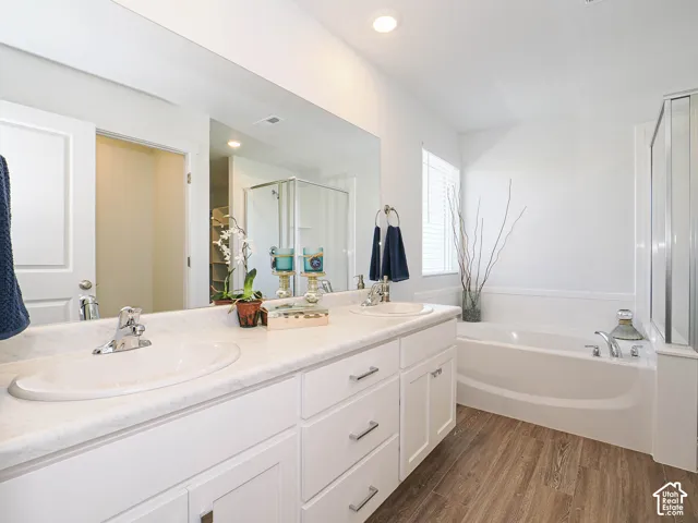 Bathroom featuring a dual-sink vanity with light-toned countertops, white cabinetry, a large wall-mounted mirror, wood-finish flooring, and a soaking tub with a glass-enclosed shower