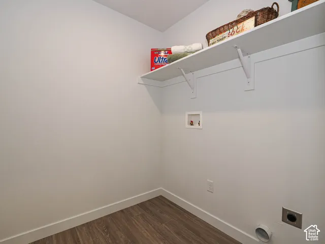 Utility area featuring wood-finish flooring, a wall-mounted shelving unit, and dedicated plumbing and electrical hookups