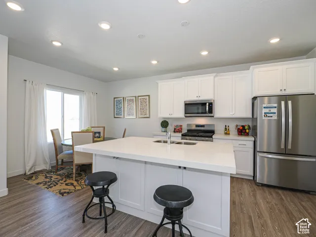 Kitchen featuring white shaker-style cabinetry, quartz countertops, a central island with an integrated sink, stainless steel appliances, and wood-finish flooring