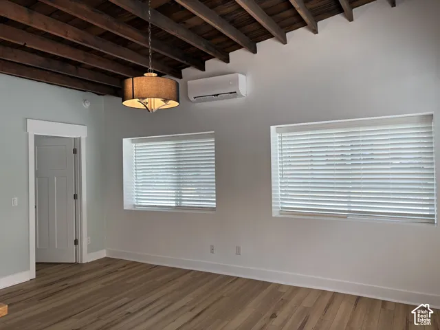 Exposed wood-beam ceiling, wood-finish flooring, two deep-set windows with horizontal blinds, wall-mounted climate control unit, and drum-style pendant light fixture