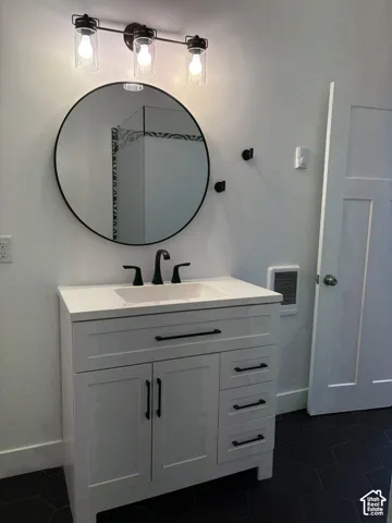 White vanity with integrated basin and matte black hardware, circular wall mirror, three-light glass fixture, and matte black plumbing fixtures set against hexagonal dark floor tiling