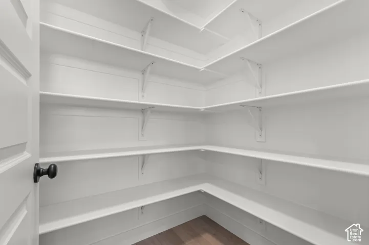 Walk-in storage space featuring wrap-around white shelving, vertical wall-mounted support brackets, and wood-finish flooring