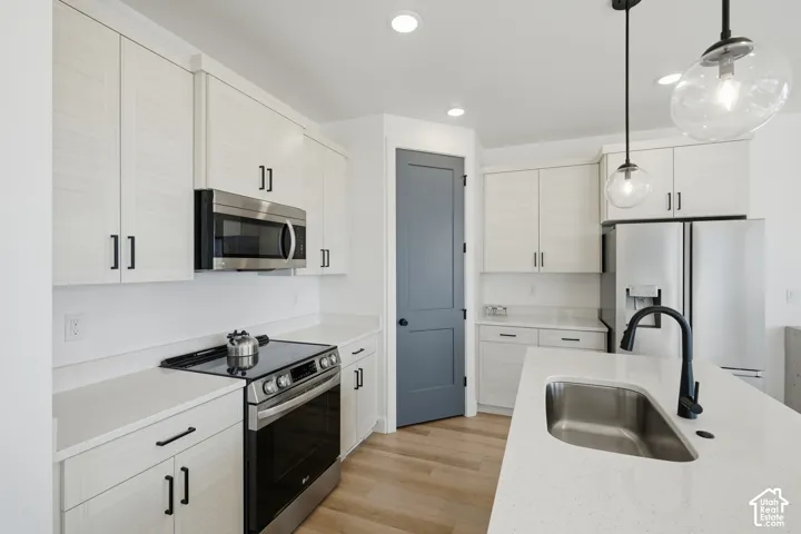 Kitchen featuring light-toned cabinetry, solid surface countertops, stainless steel appliances, a center island with an undermount sink and matte black faucet, and wood-finish flooring
