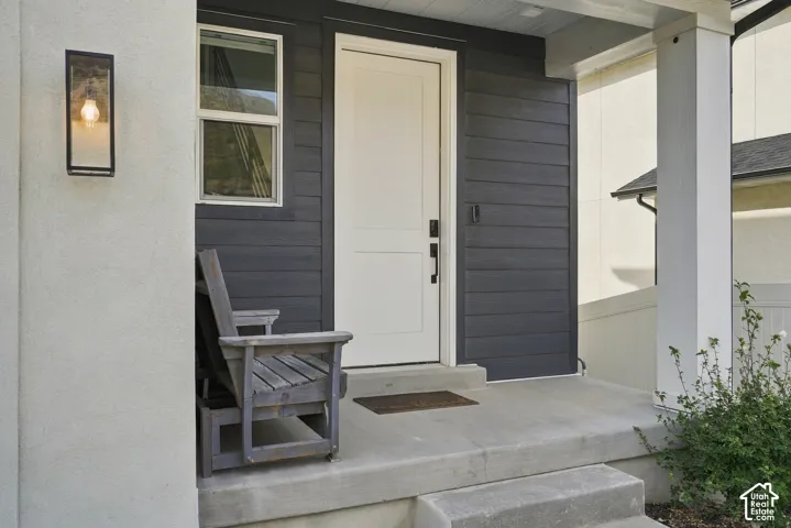 Entryway featuring dark horizontal siding, a solid panel front door, rectangular wall-mounted exterior light fixture, and a concrete porch with square support columns
