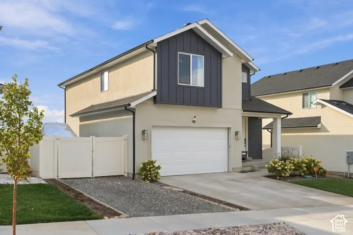 Two-story residence featuring a combination of stucco and vertical siding, concrete driveway, gravel side parking area, white vinyl fencing, and exterior wall-mounted lighting