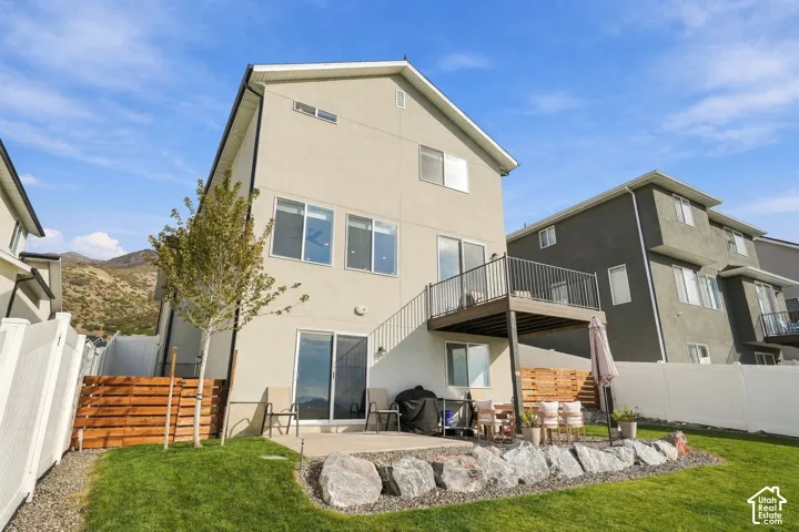 Three-story exterior with light-toned siding, elevated rear deck with metal railing, concrete patio, natural rock landscaping, and enclosed yard with wood-accent fencing