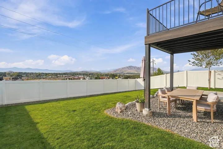 Backyard featuring a manicured lawn, white vinyl fencing, a gravel patio area, and a tiered deck with black metal railing, set against a backdrop of distant mountains