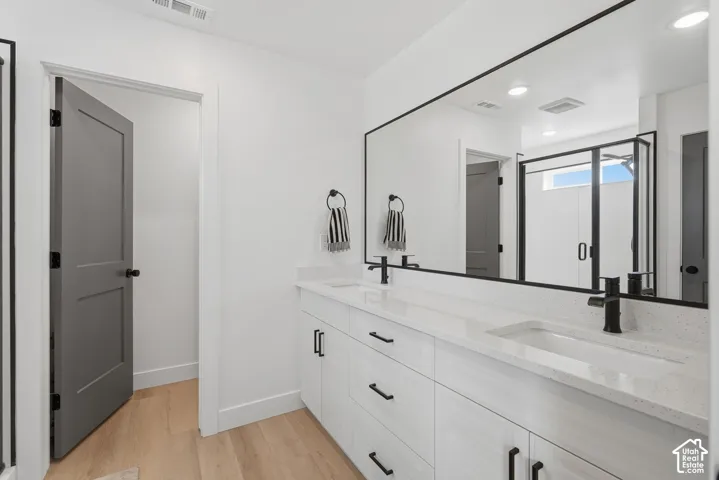 Dual-sink vanity with white cabinetry, light-toned stone-finish countertops, matte black fixtures, and a full-width mirror, complemented by wood-finish flooring and grey interior doors