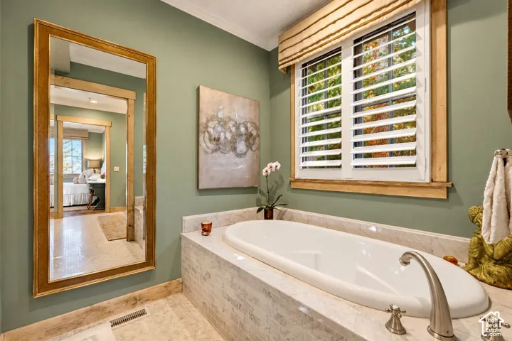 Soaking tub with a curved faucet, surrounded by stacked stone-finish tile, featuring wood-trimmed window casing, shutter-style blinds, and a large wall-mounted mirror with a wood frame