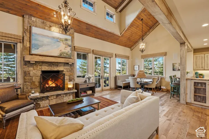 Vaulted ceiling with exposed wood beams, floor-to-ceiling stone fireplace with timber mantle, wood-finish flooring, and ornate iron chandeliers