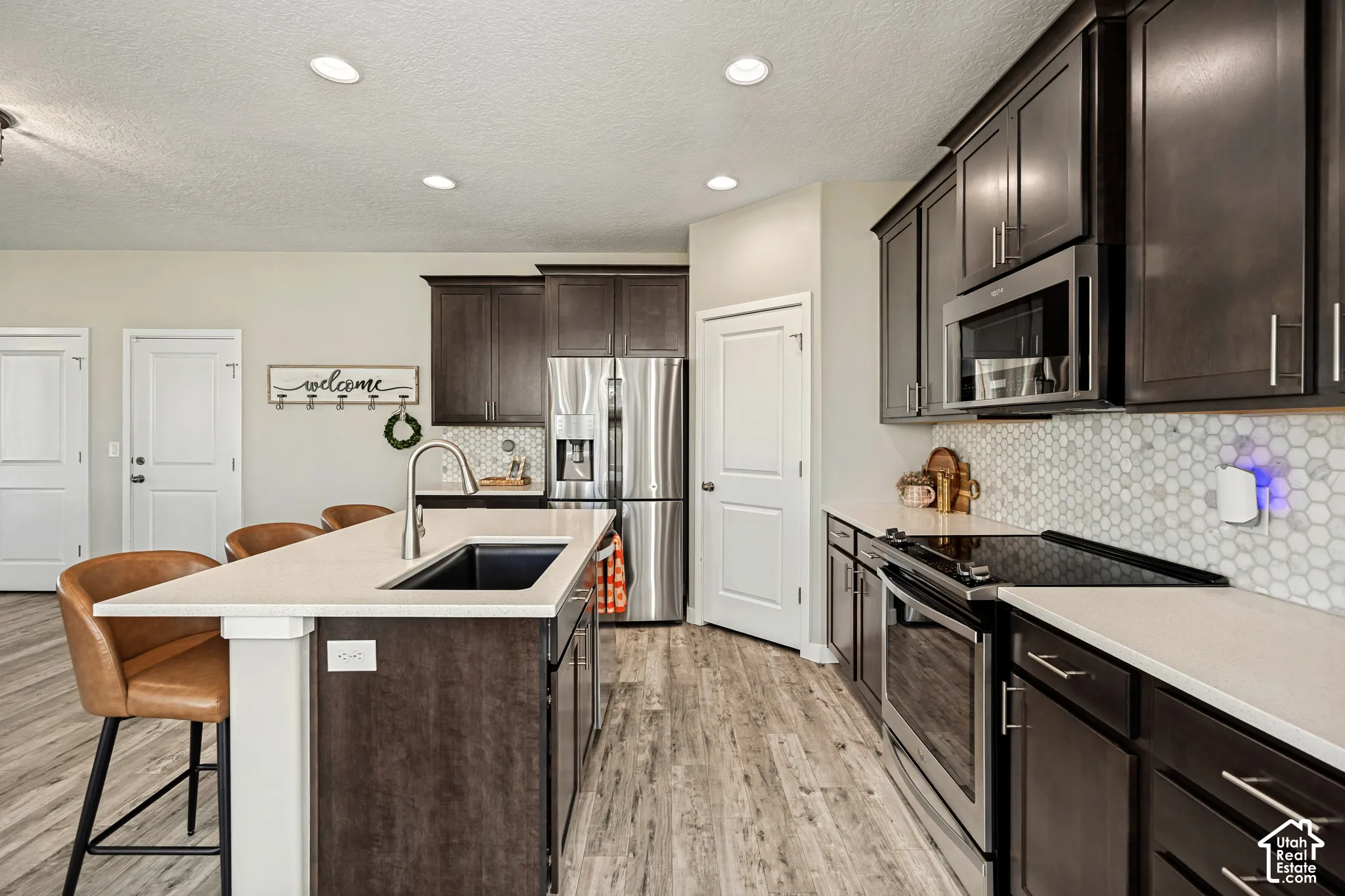 Kitchen featuring dark cabinetry, solid-surface countertops, a center island with a drop-in sink, stainless steel appliances, and a hexagonal tile backsplash