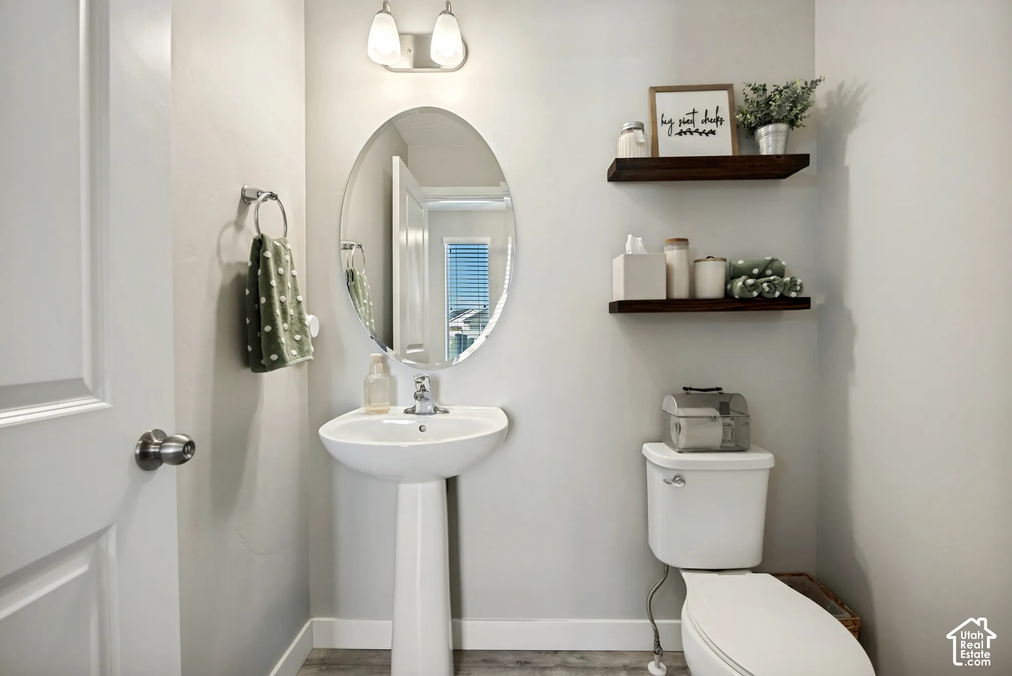 Pedestal sink with a polished chrome faucet, oval frameless mirror, dual-light vanity fixture, and two wood-finish floating shelves