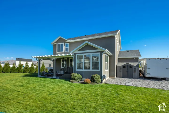 Two-story exterior featuring a stucco facade, covered rear pergola, expansive lawn, stone-surfaced side yard, and a detached storage structure