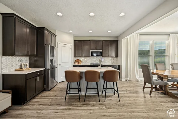 Kitchen featuring dark-finish cabinetry, a center island with countertop seating, stainless steel appliances, a hexagonal tile backsplash, and wood-finish flooring