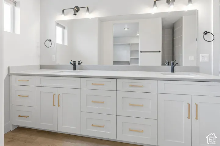 Double vanity featuring white shaker-style cabinetry, gold-tone hardware, a neutral solid-surface countertop, dual undermount sinks with matte black faucets, and a full-width mirror