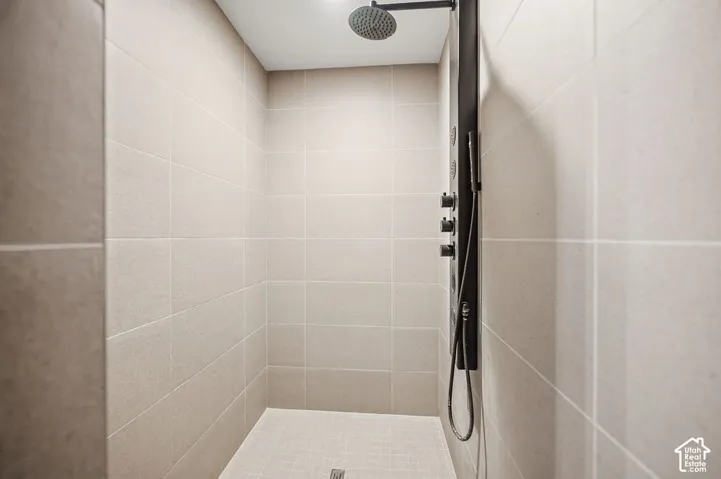 Shower stall featuring light-toned rectangular wall tile, a matte black multi-function shower panel with integrated rainfall showerhead and handheld sprayer, and square floor tiling