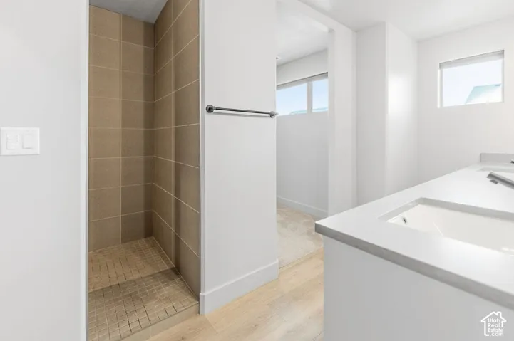 Walk-in shower with square neutral-toned wall tiling and mosaic floor tiling, dual-sink vanity with light-toned solid surface countertop, wood-finish flooring, and horizontal windows