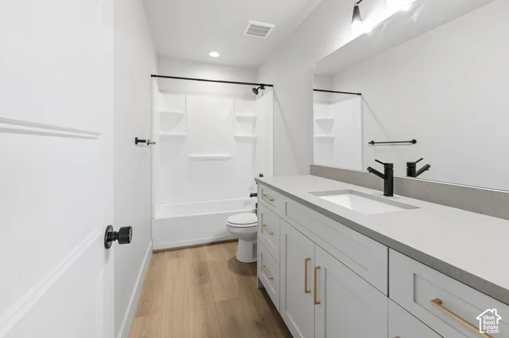 Full-width vanity with solid-surface countertop and integrated sink, matte black hardware, white shaker-style cabinetry with gold-tone pulls, tub-shower combination with built-in shelving, and wood-finish flooring