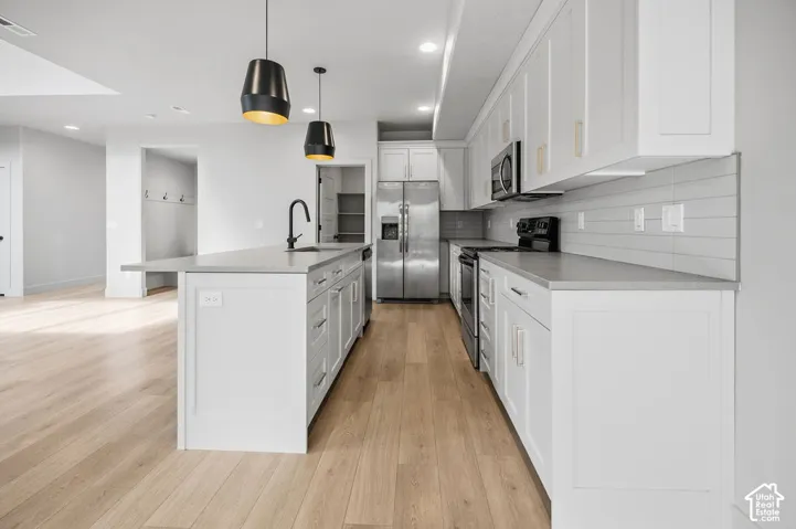 Kitchen featuring white shaker-style cabinetry, quartz countertops, a central island with an integrated sink, horizontal tile backsplash, and wood-finish flooring