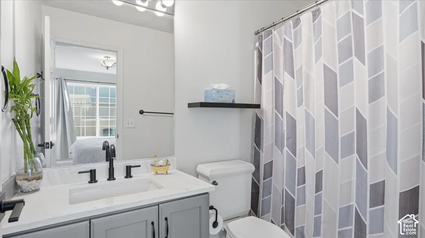 Bathroom vanity featuring a light-toned stone countertop, rectangular undermount sink, matte black widespread faucet, gray shaker-style cabinetry, and wall-mounted floating shelf