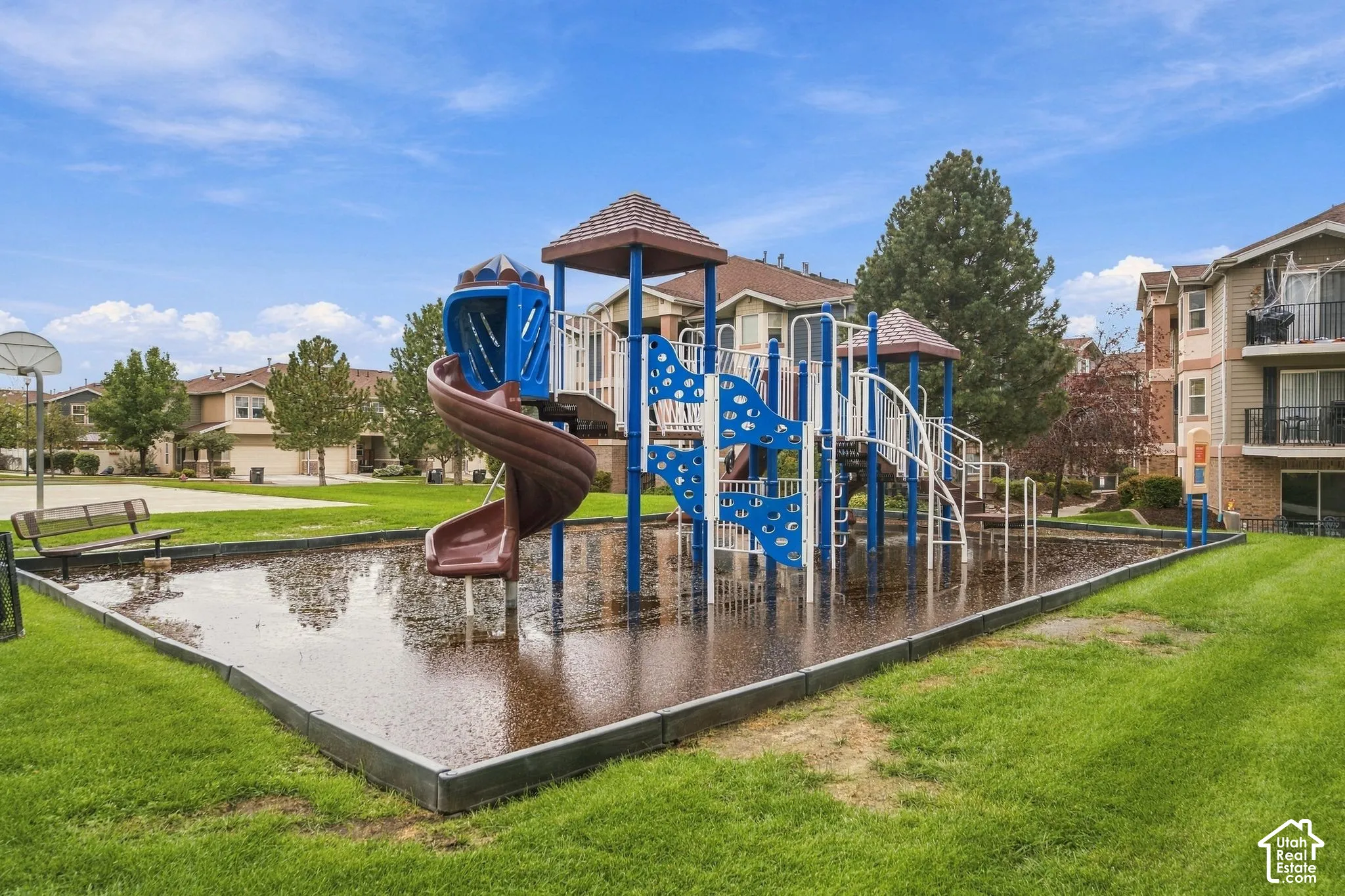 Recreational area featuring a multi-level playground structure with a spiral slide, surrounding manicured lawn, and fixed metal bench seating