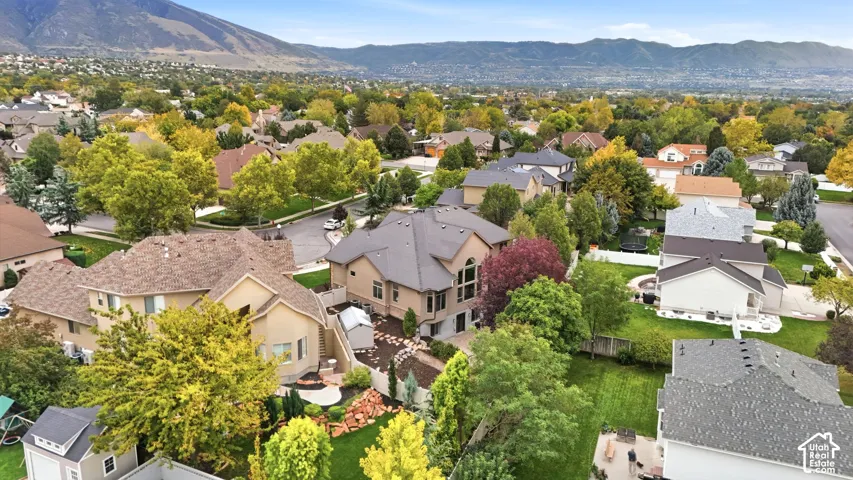 Aerial perspective of a multi-level residence featuring a multi-pitched roofline, stucco exterior, expansive windows, and a landscaped backyard with stone hardscaping, all set against a mountain backdrop