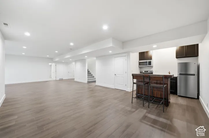 Open-concept room featuring wood-finish flooring, recessed lighting, and a kitchenette with dark-toned cabinetry, stainless steel appliances, and a built-in counter
