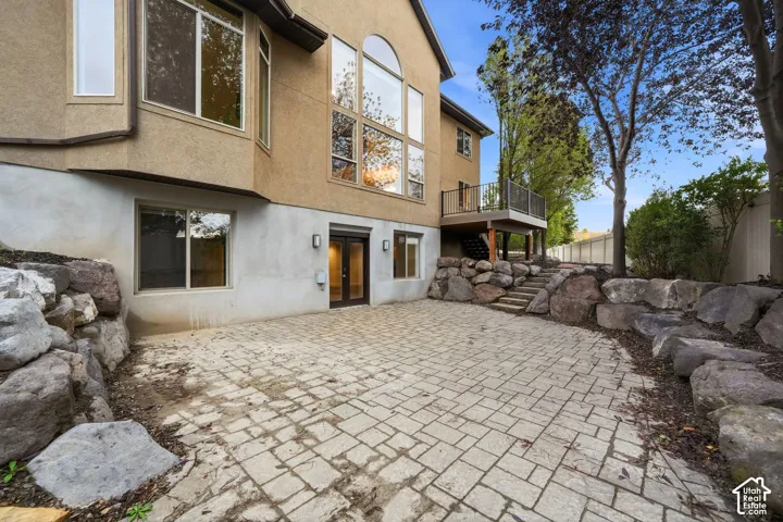 Stucco exterior featuring multi-story windows, stone retaining walls, paved stone patio, and an elevated wood-finish deck with metal railing